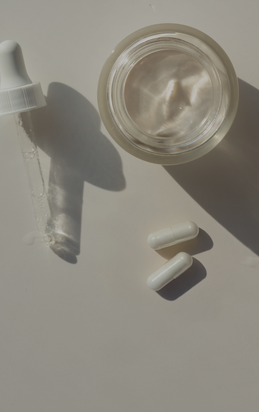 Eine transparente Glasflasche mit einem Tropfer, gefüllt mit Flüssigkeit, sowie zwei weiße Kapseln auf einer hellen Oberfläche. Die Flasche und die Kapseln werfen Schatten, wodurch eine stilvolle und moderne Präsentation entsteht.